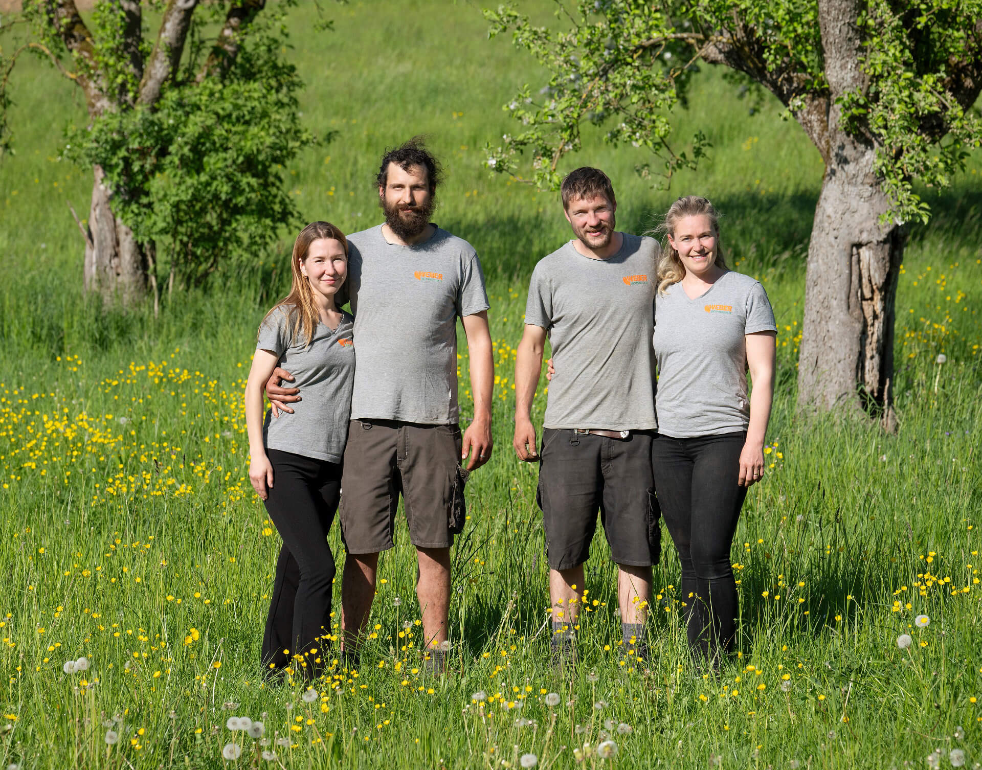 Daria Neukomm, Philipp Neukomm, Miro Weber und Tabea Weber in grauen T-Shirts stehen gemeinsam auf einer blühenden Wiese zwischen Bäumen