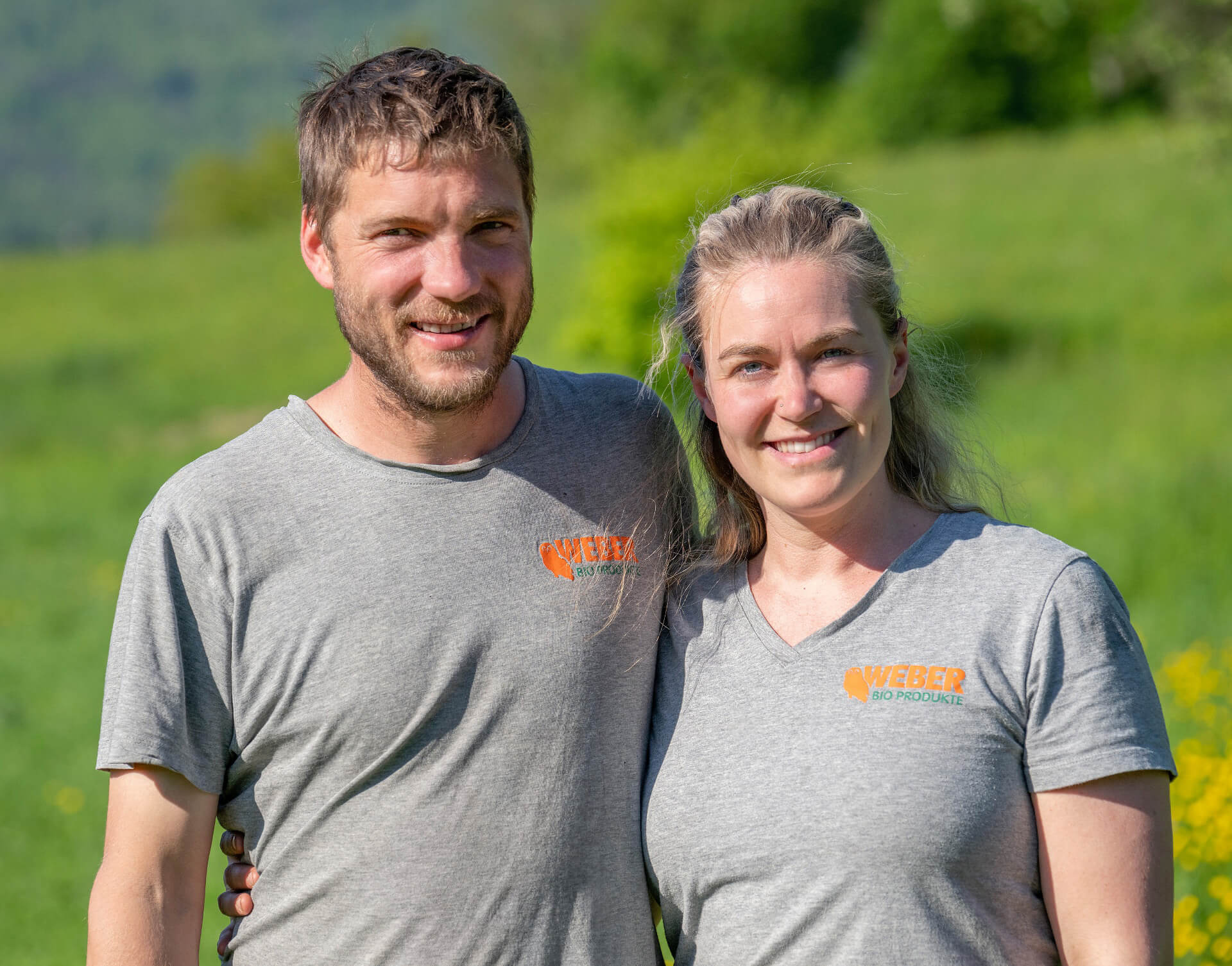 Miro Weber und Tabea Weber stehen im Grünen und tragen graue T-Shirts mit dem Aufdruck „WEBER BIO PRODUKTE“